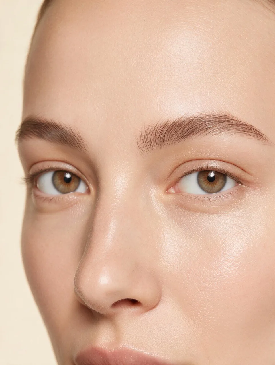Close-up of the upper half of a face with full natural eyebrows and calm eyes against a cream backdrop.